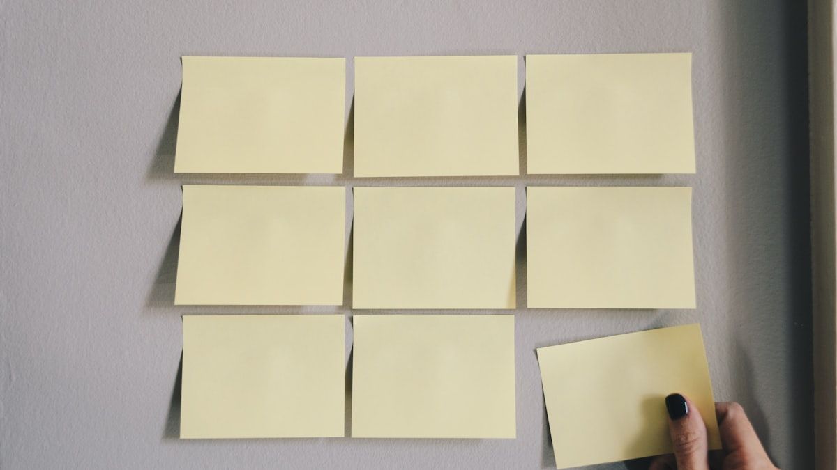 壁に並べた付箋に1枚を足そうとしている手。工程ごとに分ける場面のイメージ