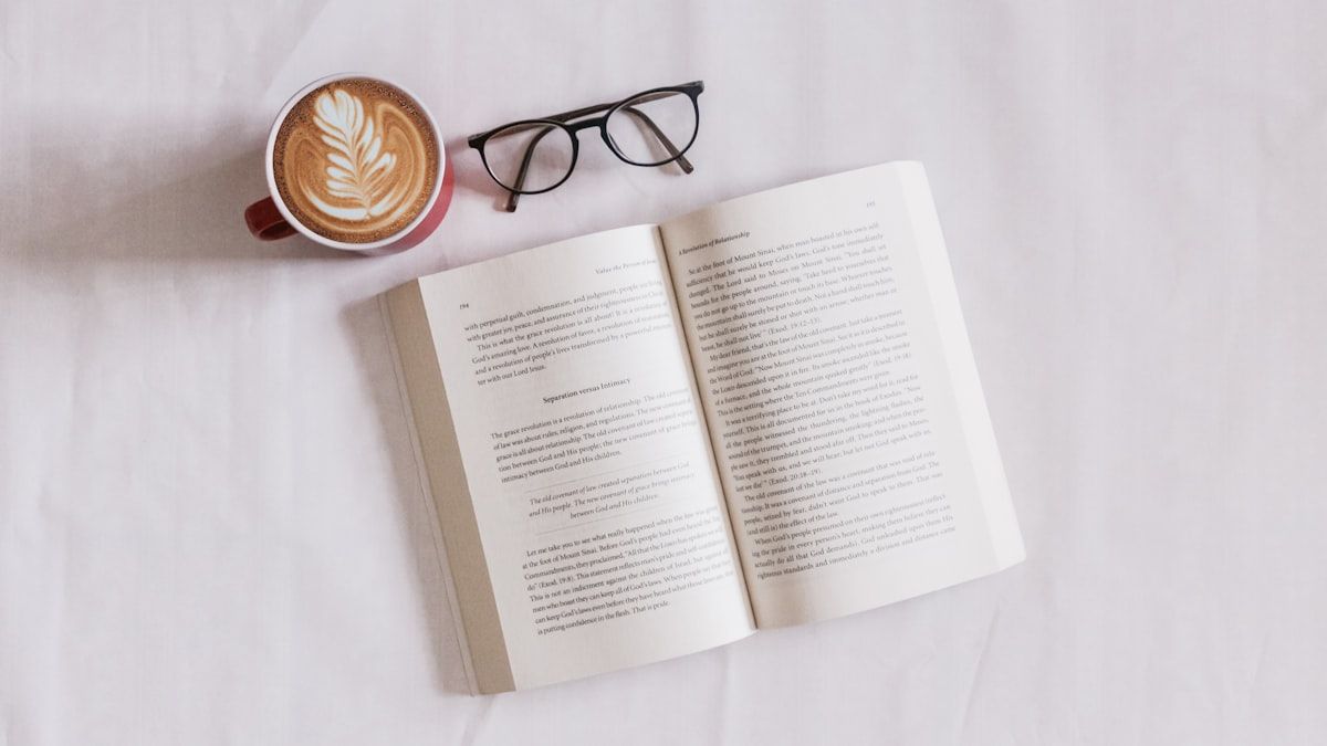 白い布の上に開かれた本と眼鏡、カフェラテの入ったカップ。細かい注記を読む場面のイメージ