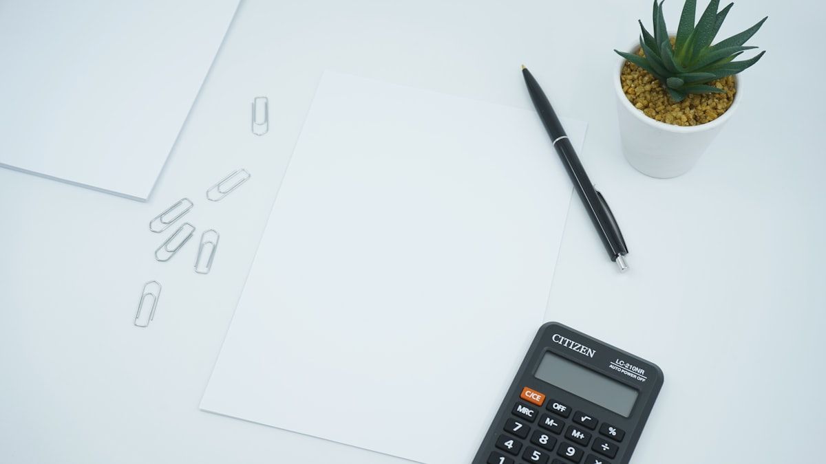 白い机に並べた白紙とクリップ、ペン、小さな電卓と観葉植物